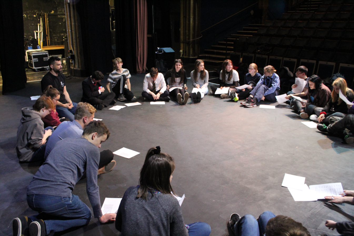 Teenagers are sitting in a circle on the stage looking at a script
