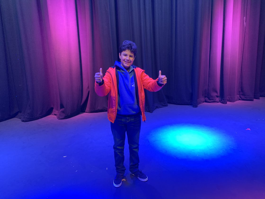 A boy stands under coloured stage lights with his thumbs up