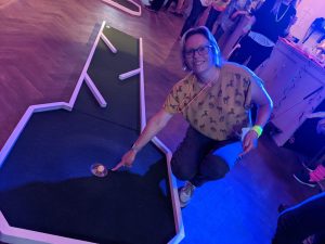 A photograph from our corporate Crazy Golf night. A guest leans down and points at a golf ball in the hole of a mini golf run.